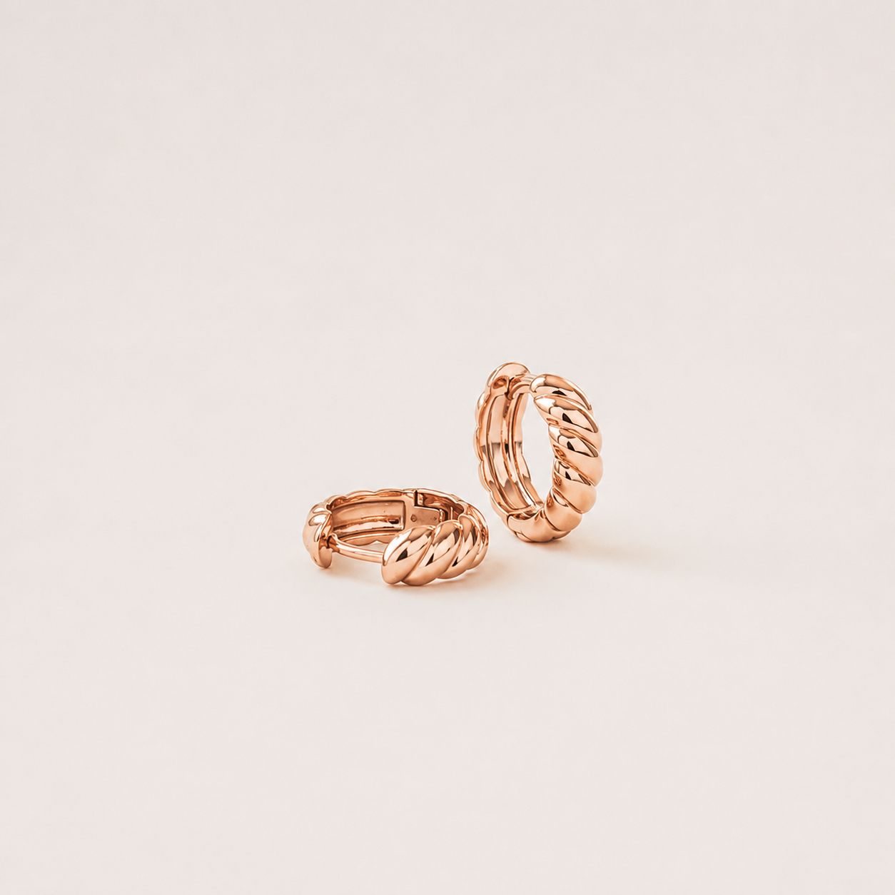 A pair of small twisted rope-style hoop earrings in polished rose gold, photographed on a clean warm off-white background.